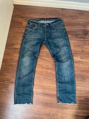 Blå jeans från Lee med råa kanter - Snygga blå jeans från Lee med klassisk femficksdesign och råa, avklippta benslut. Jeansen har en något tvättad look och Lee-logga på bakfickan. Perfekta för en avslappnad och trendig stil.
