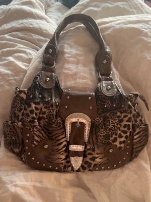 Brun handväska med djurmönster - Cool handväska med leopard- och tigermönster i bruna nyanser. Väskan har detaljer i brunt konstläder, silverfärgade nitar och en stor dekorativ spänne framtill med glittrande stenar. 