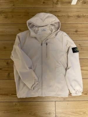 Beige vindjacka från Stone Island - Snygg beige vindjacka från Stone Island med huva och dragkedja framtill. Jackan har en bröstficka med dragkedja och den klassiska Stone Island-loggan på vänster ärm. Materialet är lätt och passar perfekt för blåsiga dagar. Står storlek S men är storlek M