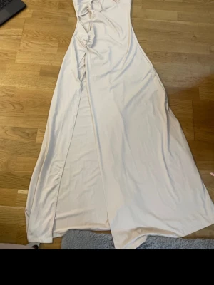 Ljus långklänning med knyt och slits - Säljer en elegant ljus långklänning med knyt i axelbanden och snygga rynkade detaljer framtill. Klänningen har en slits framtill och är tillverkad i ett mjukt, följsamt material. Perfekt för sommarens alla tillfällen.