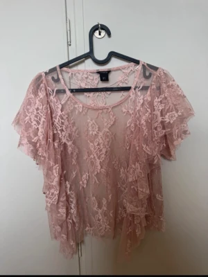 Rosa spetsblus med volanger - Söt ljusrosa blus från Lindex i transparent spets med blommigt mönster. Blusen har korta volangärmar och omlottknytning framtill. Perfekt för en trendig och romantisk look. Slutsåld💕