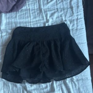 Svart kortkjol från Gina Tricot - Säljer en svart kortkjol från Gina Tricot med små glittriga prickar och volangdetaljer. Kjolen har en bred resår i midjan och är perfekt för dig som gillar en trendig och lekfull stil. Passar dig som vill ha något snyggt och bekvämt.