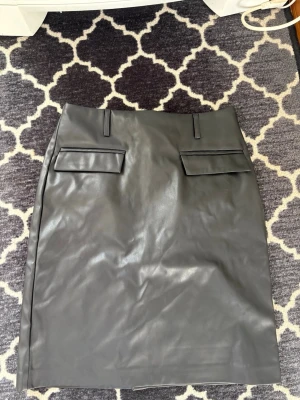 Svart pennkjol i skinnimitation - Säljer en svart pennkjol i skinnimitation med snygga fickdetaljer framtill och dragkedja bak. Kjolen har en klassisk, rak form och passar perfekt till en trendig och stilren look. Midjehöjd med hällor för bälte.