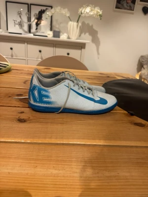 Nike vita och blå fotbollsskor - Säljer ett par vita och blå fotbollsskor från Nike med snörning och låg profil. Skorna har en blå sula och tydliga Nike-loggor på sidan och sulan. Perfekta för inomhusspel och har en sportig look.