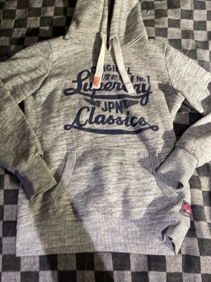 Superdry hoodie grå - Snygg hoodie från superdry. Smutsgrå färg med. Ferfekt till sommarn. Snören genom luvan med den nostalgiska orangea lappen på ena snöret