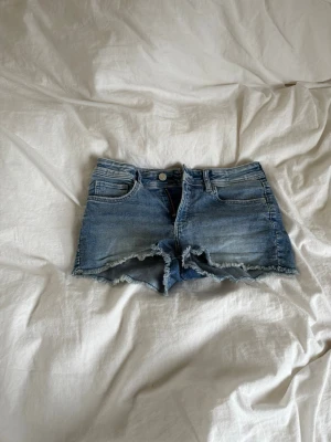 Blå denimshorts med broderad fjäril - Säljer ett par blå denimshorts med råa kanter och broderad fjäril på bakfickan. Shortsen har klassisk femficksdesign, knapp och dragkedja framtill samt slitningar. . Perfekta för sommarens och även lågmidjade med justerbar midja 