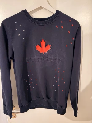 Mörkblå sweatshirt från Dsquared2 - Cool svart sweatshirt från Dsquared2 med röd lönnlöv-applikation på bröstet och diskreta färgstänk i rött och vitt. Tröjan har rund halsringning och ribbade muddar. Perfekt för dig som gillar streetwear och snygga detaljer.