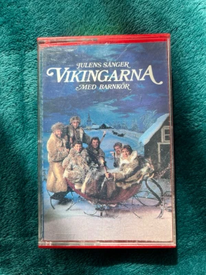 Rare old collectible Julens sånger Vikingarna kassett - Kassettband med julmusik från Vikingarna och barnkör. Omslaget har ett vintrigt motiv med släde och snö, och fodralet är rött och genomskinligt. Perfekt för dig som gillar klassisk svensk julmusik och vill ha en retro samlarpryl.