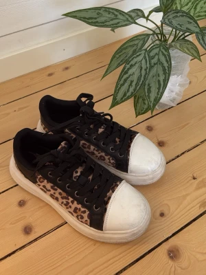 Leopardmönstrade sneakers med svarta detaljer - Snygga sneakers med leopardmönster på sidorna, svarta snören och detaljer samt vit, chunky sula och tåparti. Perfekta för dig som vill sticka ut med din stil. Ovandelen är i textil och syntet, sulan är i gummi.
