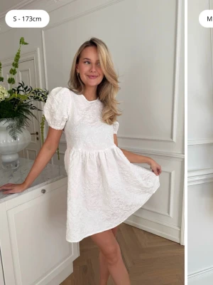 Vit kort klänning med puffärm - Superfin vit kort klänning med puffärmar och strukturmönster i tyget. Klänningen har rund halsringning och en lätt utställd kjol som ger en söt och trendig look. Helt oanvänd! 