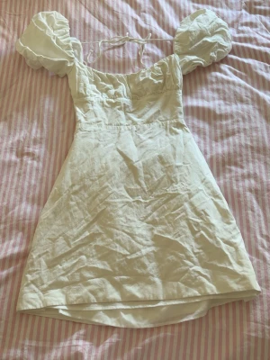 Vit kort klänning med puffärmar - Supersöt vit kort klänning med puffiga ärmar och snörning i ryggen. Klänningen har en feminin passform och är gjord i ett lätt material som passar perfekt till varma dagar. Snygg fyrkantig urringning och romantisk känsla.
