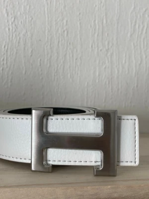 Vitt skärp med H-spänne  - Stilrent vitt skärp från med klassiskt H-format spänne i silverfärgad metall. Skärpet är gjort i skinn och har diskreta vita sömmar längs kanterna. Perfekt accessoar för att lyfta din outfit med en lyxig touch.