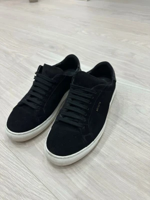 Axel Arigato - Snygga svarta sneakers från Axel Arigato i mocka med vita sulor och svarta skosnören. Diskret guldfärgad logga på sidan och stilren design som passar till det mesta. Perfekta för dig som gillar minimalistisk och trendig stil.