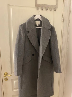 Grå lång kappa från H&M - Snygg grå kappa från H&M med klassisk rak passform Kappan har två svarta knappar framtill och stora fickor på sidorna. Materialet är ullblandning som håller dig varm under kyliga dagar. Perfekt för dig som gillar stilrena och tidlösa plagg. Passar S&M i storlek helt ny och oanvänd 