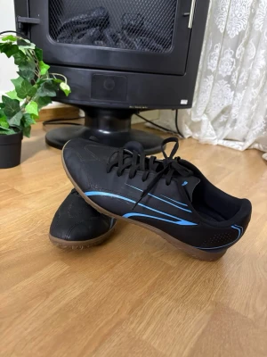 Svarta fotbollsskor med blå detaljer - Säljer ett par svarta fotbollsskor från Puma med snygga blå detaljer på sidorna och klassisk snörning. Skorna har en låg profil och gummisula som passar perfekt för inomhusspel. Materialet är syntetiskt och designen är sportig och modern.