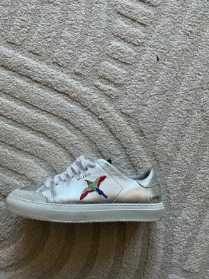 Arigato 90 fågel - Snygga vita sneakers i läder från Axel Arigato med färgglad broderad fågel på sidan och textdetaljer. Klassisk låg modell med vita skosnören och gummisula. Perfekta för en clean och trendig look.