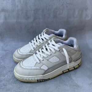 Axel Arigato skor - Snygga vita sneakers från Axel Arigato med perforerade detaljer på tån och sidan, samt robust platt sula i beige ton. Skon har snörning och är tillverkad i skinn och textil. Perfekt för dig som gillar stilrena och trendiga sneakers med låg profil. Liten defekt på insidan hälen men annars bra skick