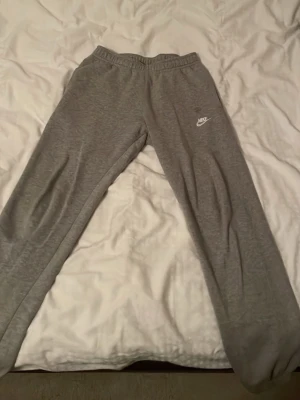 Grå Nike mjukisbyxor med logga - Säljer ett par grå mjukisbyxor från Nike med broderad vit logga på vänster ben. Byxorna har elastisk midja och raka ben, tillverkade i mjukt och bekvämt material som passar perfekt för chill eller träning.