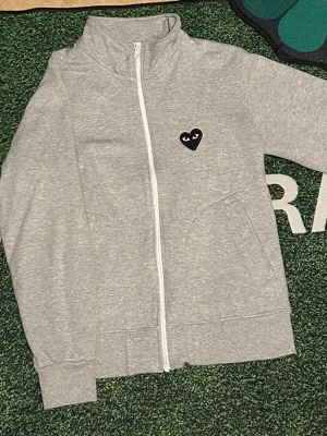 Grå hoodie från Comme des Garçons Play - Säljer en grå hoodie med vit dragkedja från Comme des Garçons Play. Den har ett svart hjärta med ögon broderat på bröstet och två fickor framtill. Materialet är mjukt och bekvämt, perfekt för en avslappnad stil.