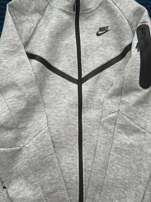 Grå Nike Tech Fleece hoodie - Snygg grå Nike Tech Fleece hoodie med hel dragkedja och svarta detaljer. Har en huva och en praktisk ficka med dragkedja på ärmen. Tillverkad i mjukt och lätt material som ger en modern look. Perfekt för dig som gillar sportig och stilren streetwear.