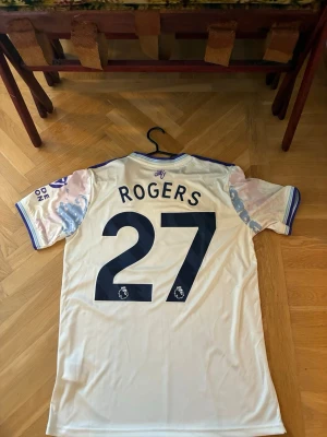 Aston Villa bortatröja Rogers 27 - Säljer en Aston Villa bortatröja från Adidas med Rogers och nummer 27 på ryggen. Tröjan är vit med lila och blå detaljer, har klubbmärke och Betano-logga på bröstet samt Premier League-märke på ärmen. Perfekt för fotbollsfans som vill sticka ut på läktaren.