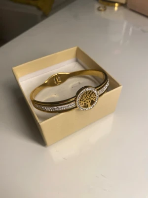 Guldigt armband med livets träd - Snyggt armband i guldfärg med ett runt hänge som har ett utskuret livets träd. Armbandet är dekorerat med små gnistrande stenar längs sidorna för extra bling. Perfekt accessoar för dig som gillar stilrena men ändå iögonfallande smycken.