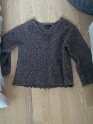 Brun stickad tröja med v-ringning - Mysig brun stickad tröja med v-ringad hals. Tröjan har en lite lös passform och är perfekt för kyliga dagar. Materialet är  en ull- eller mohairblandning som ger en fluffig känsla.