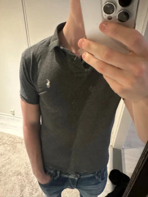 Mörkgrå pikétröja från Polo Ralph Lauren - Säljer en mörkgrå pikétröja från Polo Ralph Lauren med klassisk krage och två knappar framtill. Tröjan har korta ärmar och den ikoniska vita logotypen broderad på bröstet. Perfekt till jeans eller shorts för en clean look.