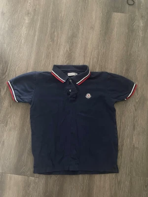 Marinblå polotopp från Moncler - Säljer en marinblå polotopp från Moncler med klassisk krage och korta ärmar. Toppens krage och ärmslut har snygga ränder i vitt och rött. Moncler-loggan är broderad på bröstet. Perfekt för dig som gillar stilrena och sportiga plagg. Storleken passar som en XS-S