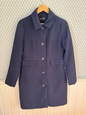 Mörkblå klassisk rock från KappAhl - Stilren mörkblå rock från KappAhl med rak passform och stora knappar framtill. Rocken har klassisk krage, långa ärmar och två dekorativa fickor. Perfekt för dig som gillar enkel och tidlös design. 