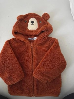 Brun teddyjacka med björnmotiv - Supermysig brun teddyjacka från Lindex med huva som har söta björnöron och broderat björngesikt. Jackan är gjord i mjukt och fluffigt teddy-material och har dragkedja framtill. Perfekt för att hålla sig varm under kyliga dagar.
