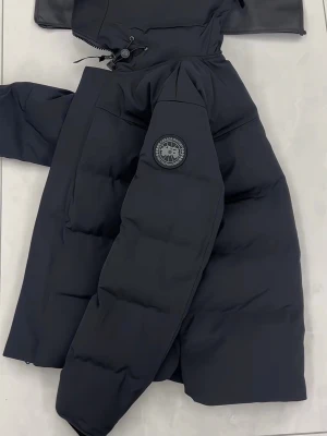 Svart pufferjacka från Canada Goose - Snygg svart pufferjacka från Canada Goose med huva och klassisk logotyp på ärmen. Jackan har en stilren design, dragkedja framtill och är tillverkad i slitstarkt material med vaddering som håller dig varm under kalla dagar.