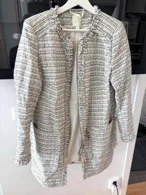 Tweedkappa - Stilren vit/ljusgrå kappa i tweed med svarta inslag och fransade kanter. Rak modell med rund halsringning, dragkedja framtill och två stora fickor. Perfekt för en trendig och chic look.      