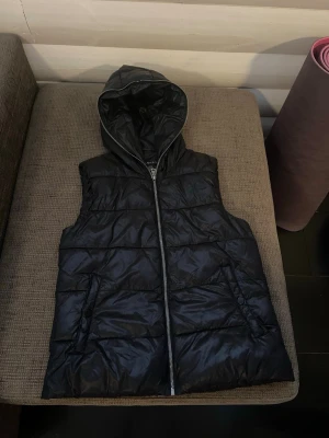 Svart dunväst  - Svart dunväst  med dragkedja framtill och två fickor med knappstängning. Västen har en stor, mysig huva och quiltad design som ger en cool look. Perfekt för lager-på-lager och chill dagar ute. Materialet är glansigt och lätt.