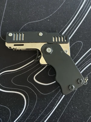 Svart och silver gummiband pistol - Unik gummiband pistol i form av en liten pistol, svart och silverfärgad med metallhandtag och detaljer i plast. Kompakt design som kan fällas ihop för enkel förvaring. Perfekt som samlarpryl eller cool accessoar till nyckelknippan.