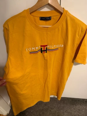 Gul t-shirt från Tommy Hilfiger - Snygg gul t-shirt från Tommy Hilfiger med broderad logga och text på bröstet. Klassisk passform och rund halsringning. Tillverkad i mjuk bomull som känns skön mot huden. Perfekt för dig som vill ha en färgstark och stilren look.