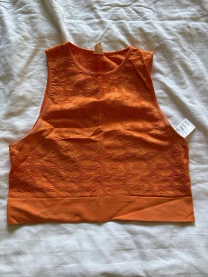 Orange ärmlös linnetopp - Snygg orange linnetopp med diskret blommönster och ribbad nederkant. Toppen är ärmlös och har en hög halsringning, perfekt för en trendig och sportig look. Materialet känns stretchigt och bekvämt, vilket gör den enkel att bära till olika outfits.