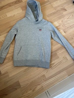 Grå hoodie från GANT med logga - Snygg grå hoodie från GANT med broderad logga och sköld på bröstet. Klassisk passform med huva och känguruficka framtill. Tillverkad i mjukt sweatshirtmaterial som är perfekt för chill dagar. Ribbstickade muddar vid ärmslut och nederkant.
