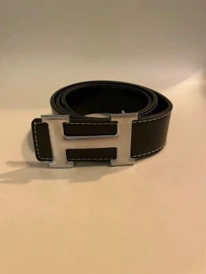 Svart skärp med H-spänne  - Snyggt svart  med ett stort H-format metallspänne i silver. Skärpet har vita kontrastsömmar längs kanterna och är gjort i slätt skinn. Perfekt accessoar för att lyfta vilken outfit som helst.