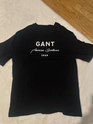 Svart GANT t-shirt med tryck - Snygg svart t-shirt från GANT med vitt tryck på bröstet där det står 'GANT American Sportswear 1949'. Klassisk passform och rund halsringning. Perfekt till jeans eller shorts för en clean och enkel stil.