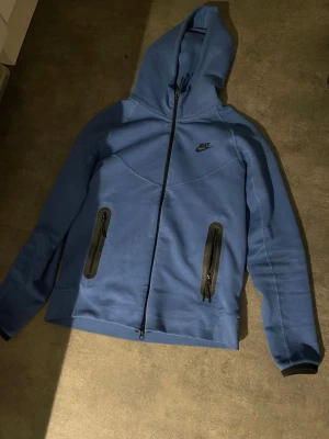 Blå  Nike tech med dragkedja - Snygg blå  Nike tech dress med hel dragkedja framtill och två svarta dragkedja eförsedda fickor. Klassisk Nike-logga på bröstet och huva med dragsko. Tillverkad i mjukt bomullsmaterial som känns skönt mot huden. Perfekt för en avslappnad och sportig stil.