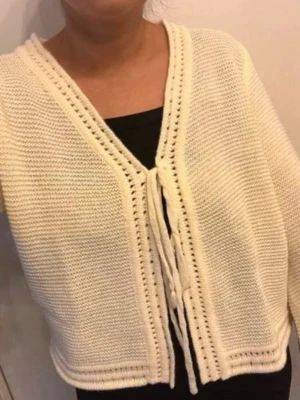 Krämvit stickad kofta med knytband - Säljer en krämvit stickad kofta med v-ringning och dekorativa knytband framtill. Koftan har breda ärmar och ett luftigt, mjukt material som känns skönt mot huden. Perfekt att slänga över en topp för en chill look.