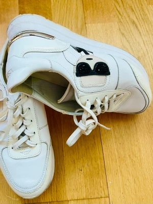 Vita sneakers Karl Lagerfeld  - Snygga vita sneakers från Karl Lagerfeld med svart broderad detalj av solglasögon på sidan. Skorna har klassisk låg modell, vita snören och är tillverkade i läder. Perfekta för dig som gillar stilrena och trendiga skor med en twist.