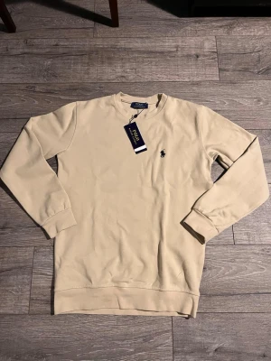 Beige sweatshirt från Polo Ralph Lauren - Stilren beige sweatshirt från Polo Ralph Lauren med klassisk rund halsringning och broderad logga på bröstet. Tröjan har långa ärmar och ribbade muddar vid ärmslut och nederkant. Perfekt för en avslappnad och snygg look.