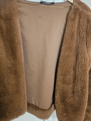 Brun pälsjacka från Gina Tricot - Mysig brun pälsjacka från Gina Tricot i fuskpäls. Jackan har en rak, kortare modell med långa ärmar och slät insida. Perfekt för att hålla värmen under kalla dagar och ger en trendig look.