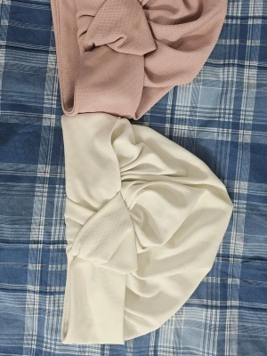 Två turbanmössor i beige och rosa - Säljer två snygga turbanmössor i mjukt tyg, en i ljusbeige och en i ljusrosa. Båda har en twistad knut framtill och är perfekta för att ge din look en trendig touch. Passar dig som gillar stilrena accessoarer.