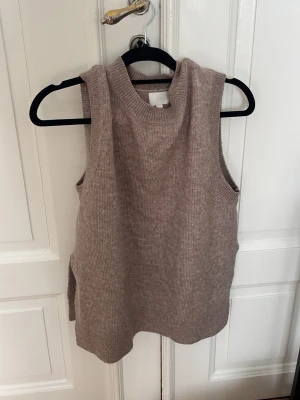 Beige stickad väst med slits - En stilren beige stickad väst med rund hals och slits i sidan. Perfekt att bära över en skjorta eller t-shirt för en trendig lager-på-lager-look. Västen har en mjuk och ribbad struktur som ger en avslappnad men snygg vibe.