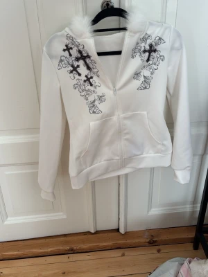 Vit hoodie med fuskpäls och brodyr - Säljer en vit hoodie med dragkedja, stor huva kantad med fluffig fuskpäls och snygg svart brodyr med kors och mönster på bröstet. Hoodien har två stora fickor fram och är gjord i mjukt material, perfekt för en trendig och bekväm look.
