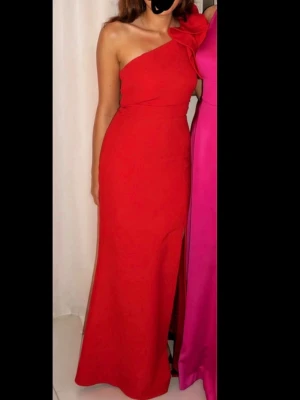 Festklänning - Snygg röd långklänning med oneshoulder-design och stor dekorativ rosett på axeln. Klänningen har en slits framtill och figurnära passform som framhäver siluetten. Perfekt för dig som vill sticka ut och känna dig extra glam. Materialet känns mjukt och bekvämt. Endast använts 1 gång och är därmed i nyskick.     Nypris 2000kr Mitt pris 1100