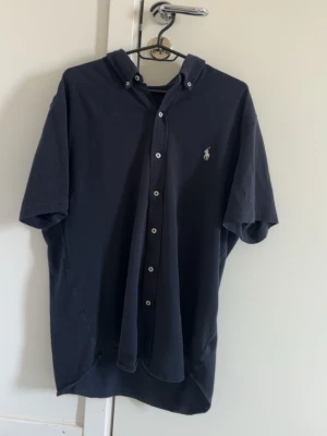 Mörkblå skjorta från Polo Ralph Lauren - Snygg mörkblå skjorta från Polo Ralph Lauren i featherweight mesh. Skjortan har korta ärmar, klassisk krage med knappar och den ikoniska loggan broderad på bröstet. Perfekt för en clean och stilren look.